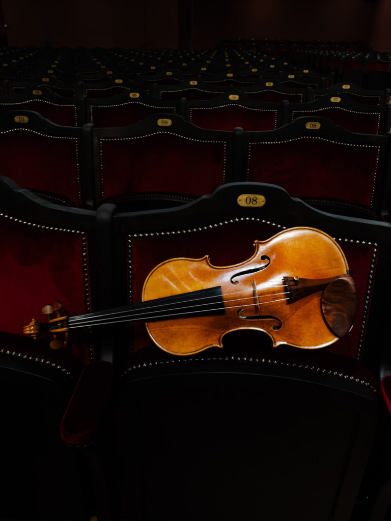 A violin rests across the back of a red velvet theater seat inside an empty auditorium filled with rows of matching chairs. Low, directional light highlights the warm wood of the instrument against the dark surroundings, creating a quiet and contemplative atmosphere.