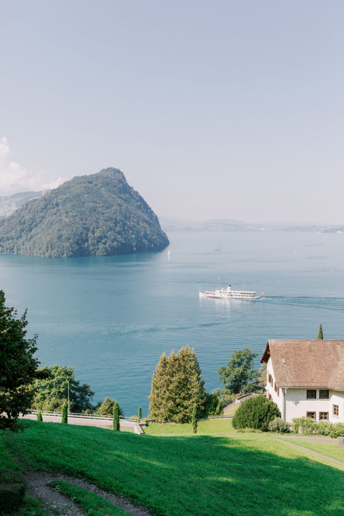 A passenger boat crosses a calm lake beneath a forested hill, viewed from an elevated hillside overlooking the water. Soft morning light illuminates the landscape, with green lawns, trees, and lakeside buildings framing the scene.