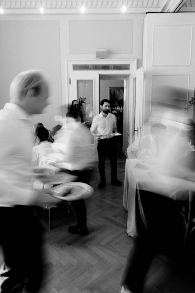 Guests and staff move through a wedding reception dining room, with motion blur capturing the pace of service and conversation. The black-and-white image contrasts blurred figures with a server standing in a doorway, while tables and architectural details define the interior space.