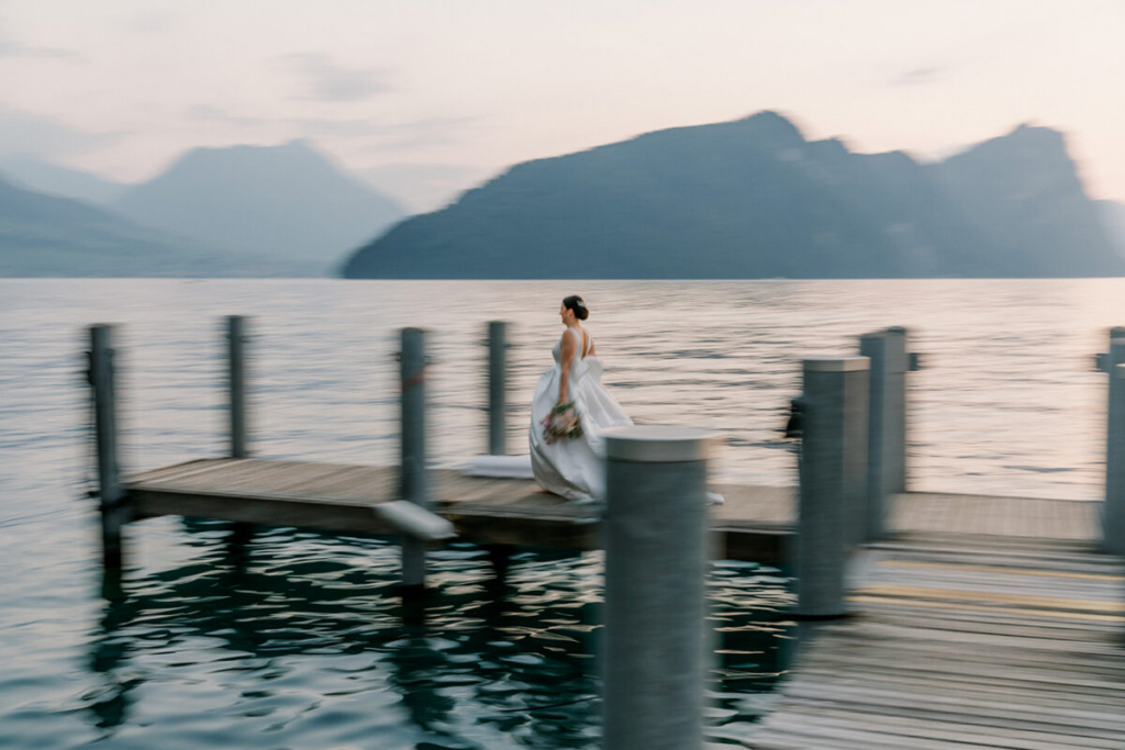A bride in a white wedding gown walks along a lakeside dock at dusk, holding a bouquet as calm water and distant mountains fill the background. The image uses intentional motion blur, creating a sense of movement while soft pastel light softens the scene and reflects across the lake.