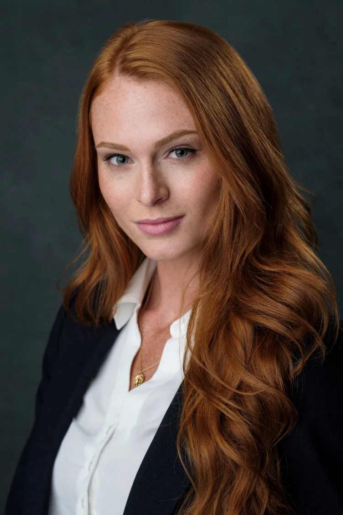 A close-up studio portrait of a woman with long red hair looking directly at the camera. She wears a white blouse under a dark blazer, with soft lighting and a dark gray background.