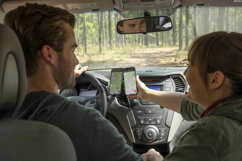 A man drives a car along a forest road while a woman in the front passenger seat holds up a smartphone showing a navigation map. Another phone displaying a map is mounted on the dashboard, and trees are visible through the windshield.