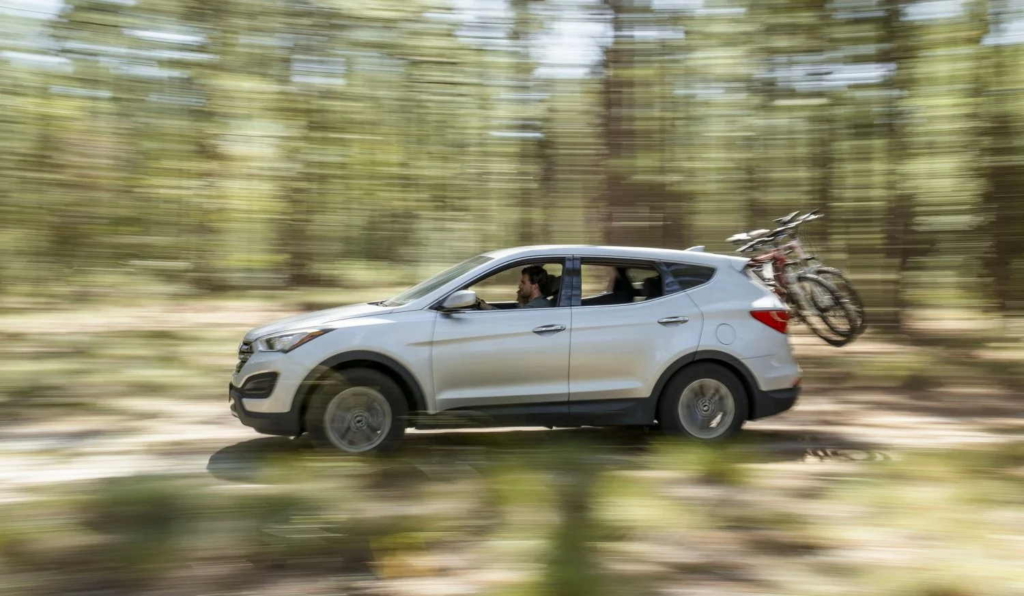 A white SUV drives along a forest road with two bicycles mounted on a rear bike rack. Motion blur in the trees and ground emphasizes the vehicle's movement through the wooded landscape.
