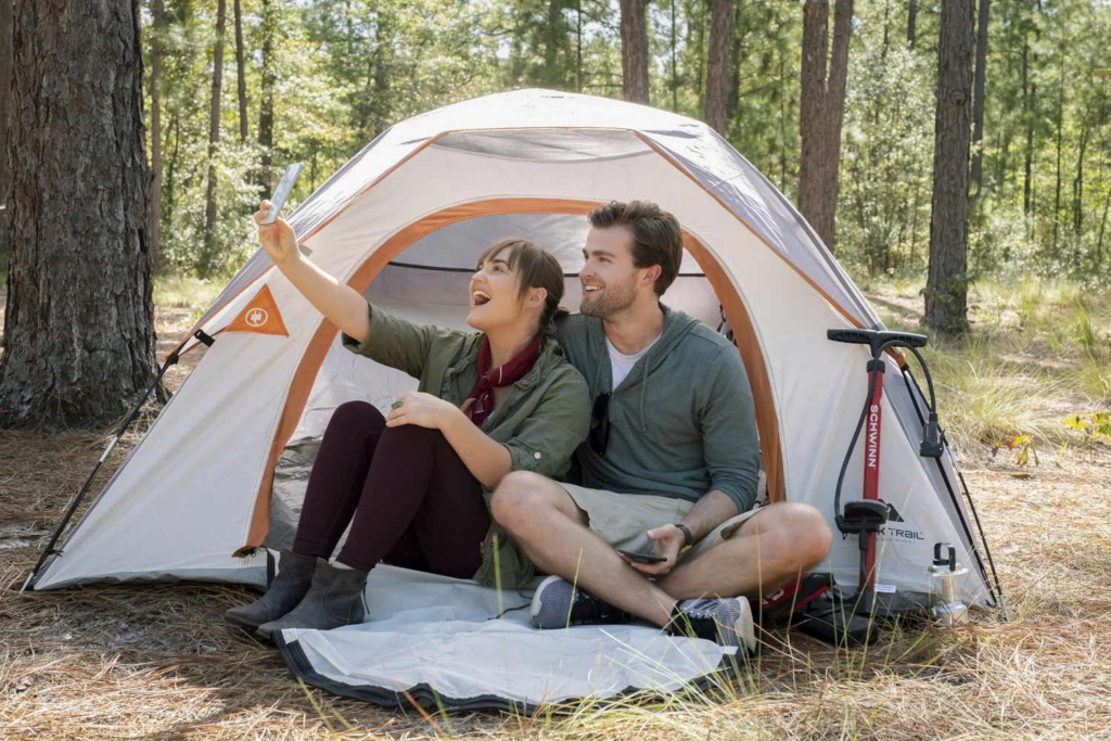 A man and a woman sit together at the entrance of a camping tent in a forest, smiling as the woman holds up a smartphone. Sunlight filters through the trees, with camping gear and a bicycle pump beside the tent.