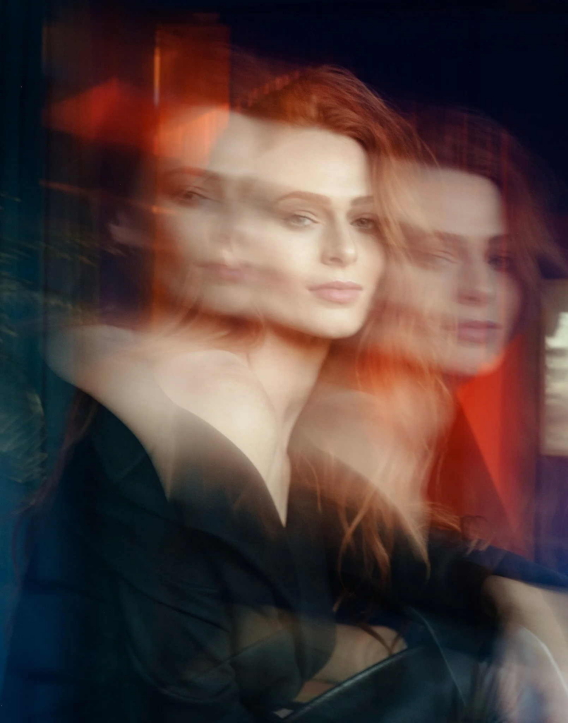 A woman with long red hair wears a black off-the-shoulder outfit and looks calmly toward the camera. Motion blur and reflections create several overlapping views of her face, while soft, warm light gives the portrait a dreamy, layered effect.