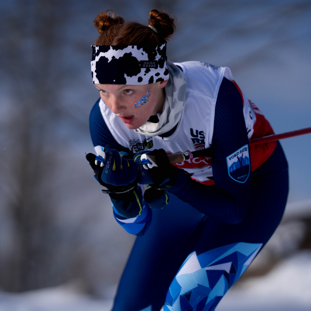 A cross-country skier races uphill through a snowy course, leaning forward with both ski poles planted as she drives through the climb. Her focused expression, patterned headband, and blue race suit stand out against a softly blurred winter forest background.