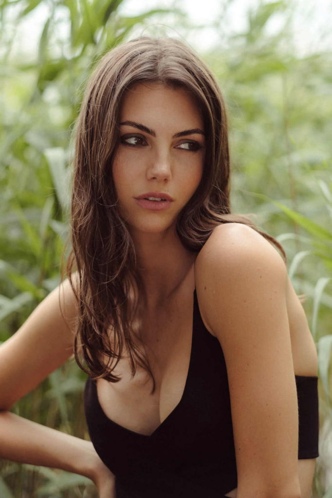 A woman with shoulder-length dark hair wears a black sleeveless top and sits among tall green plants outdoors. She looks to the side with a calm expression, while soft natural light and the blurred greenery create a relaxed, natural setting.