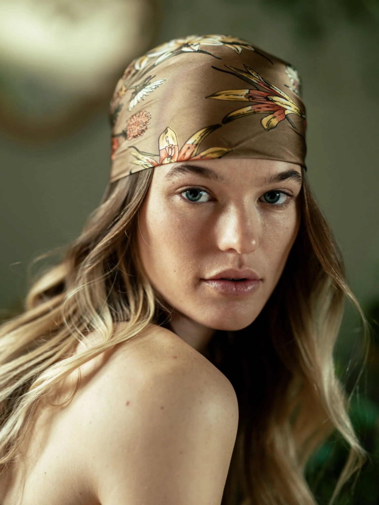 A close-up portrait of a woman with long blonde hair wearing a floral silk headscarf. She looks directly at the camera with a calm expression, while soft natural light and a blurred green background create a warm, peaceful atmosphere.