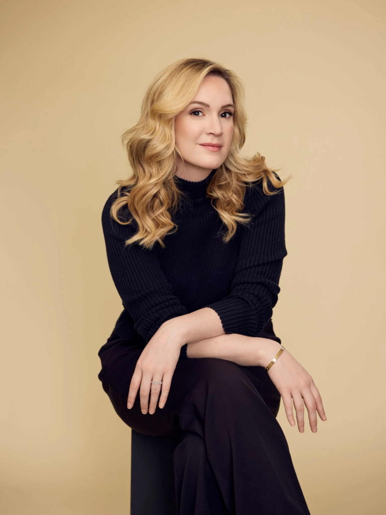 A woman with long blonde hair sits on a stool against a plain beige background, looking toward the camera with a relaxed expression. She wears a black sweater and dark pants, with her hands resting naturally in her lap under soft, even lighting.