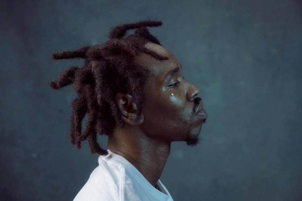 A side-profile portrait of a person with short dreadlocks looking straight ahead against a dark gray background. They wear a white shirt, and soft light highlights the contours of their face and hair.