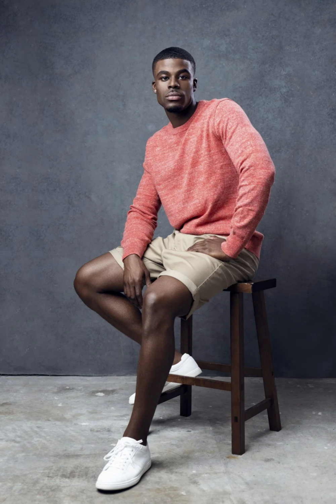 A man sits on a wooden stool in a studio, looking directly at the camera. He wears a coral-colored sweater, beige shorts, and white sneakers, with a textured gray background softly lit behind him.