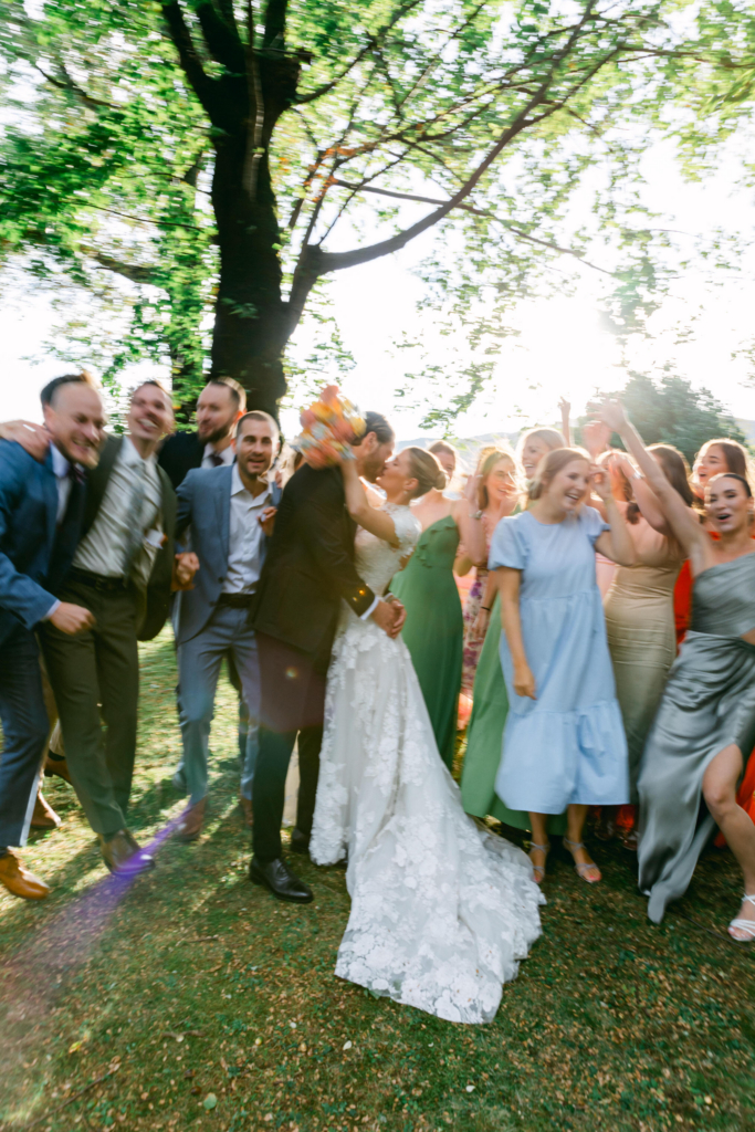 A bride and groom kiss in the center of a group of wedding guests gathered beneath a large tree during an outdoor celebration. Bright sunlight filters through the leaves while motion blur and raised arms capture the movement and energy of the moment.